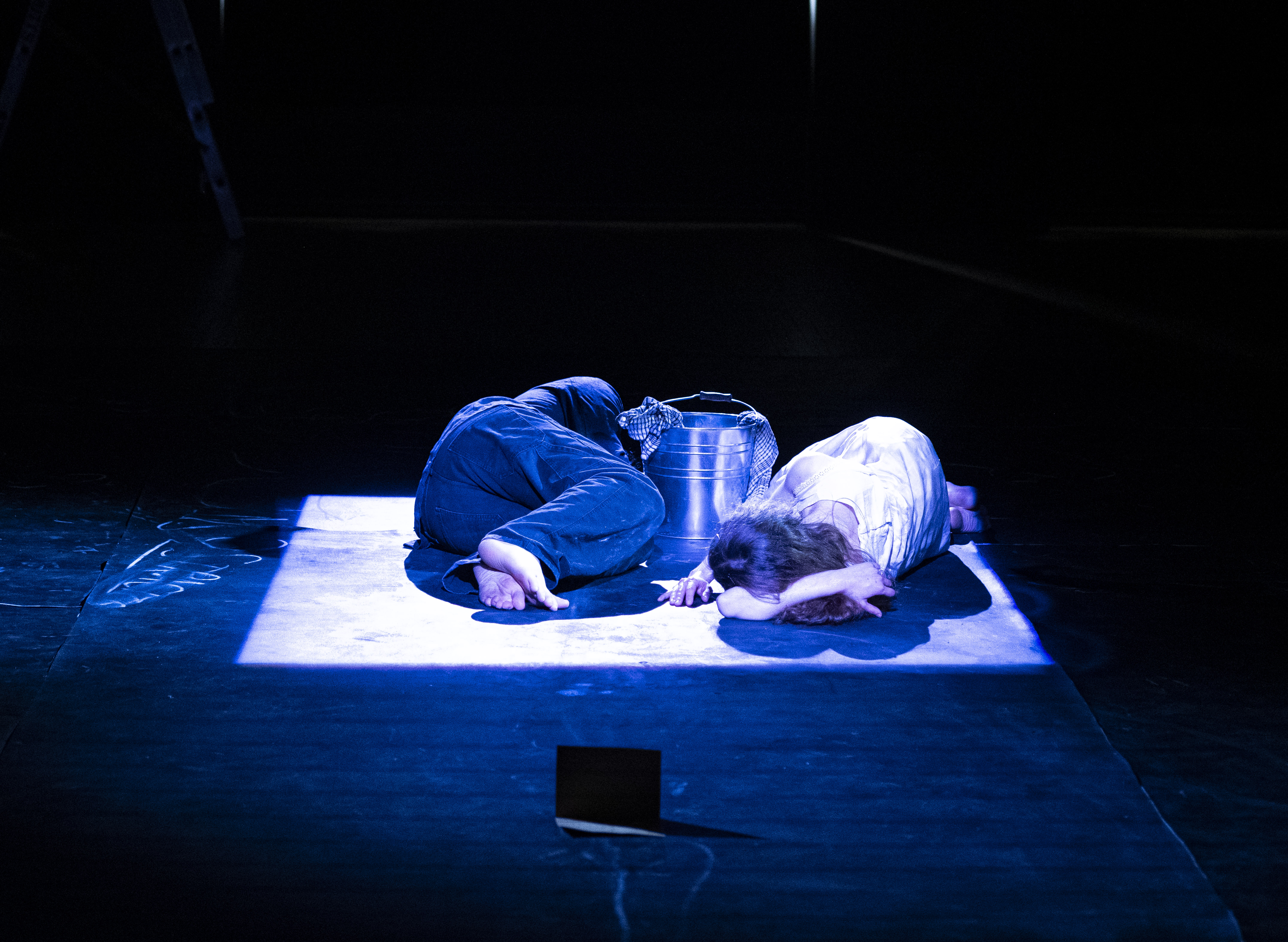 Two figures curled together in a lit square on a dark stage.