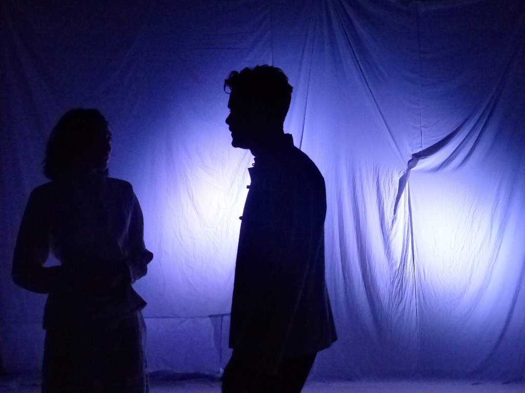 Two figures in silhouette facing each other against a violet-lit sheet.