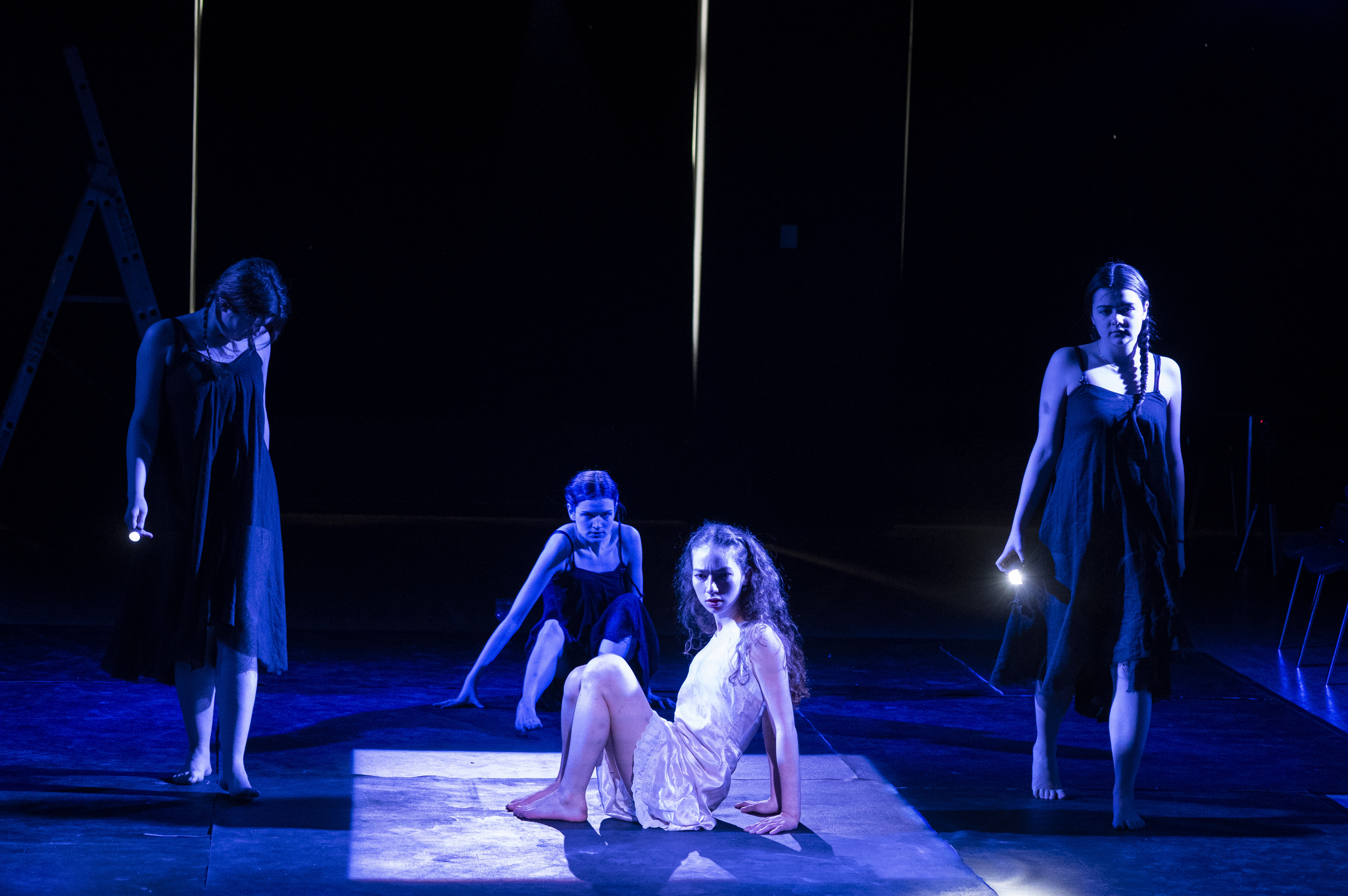 Eurydice seated in a lit square as two of the Stones cross the stage.