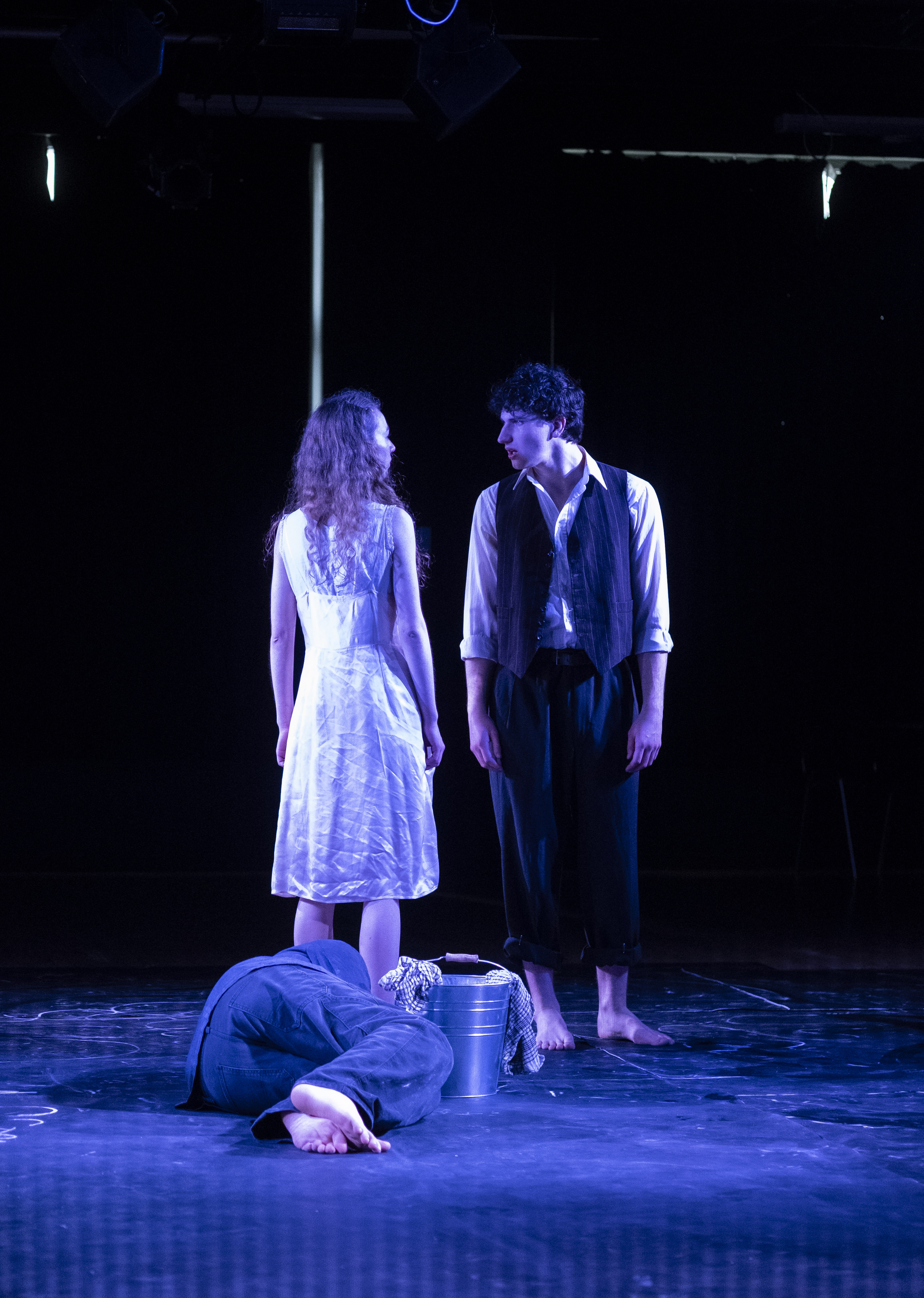 Two figures standing over a curled body beside a metal bucket.