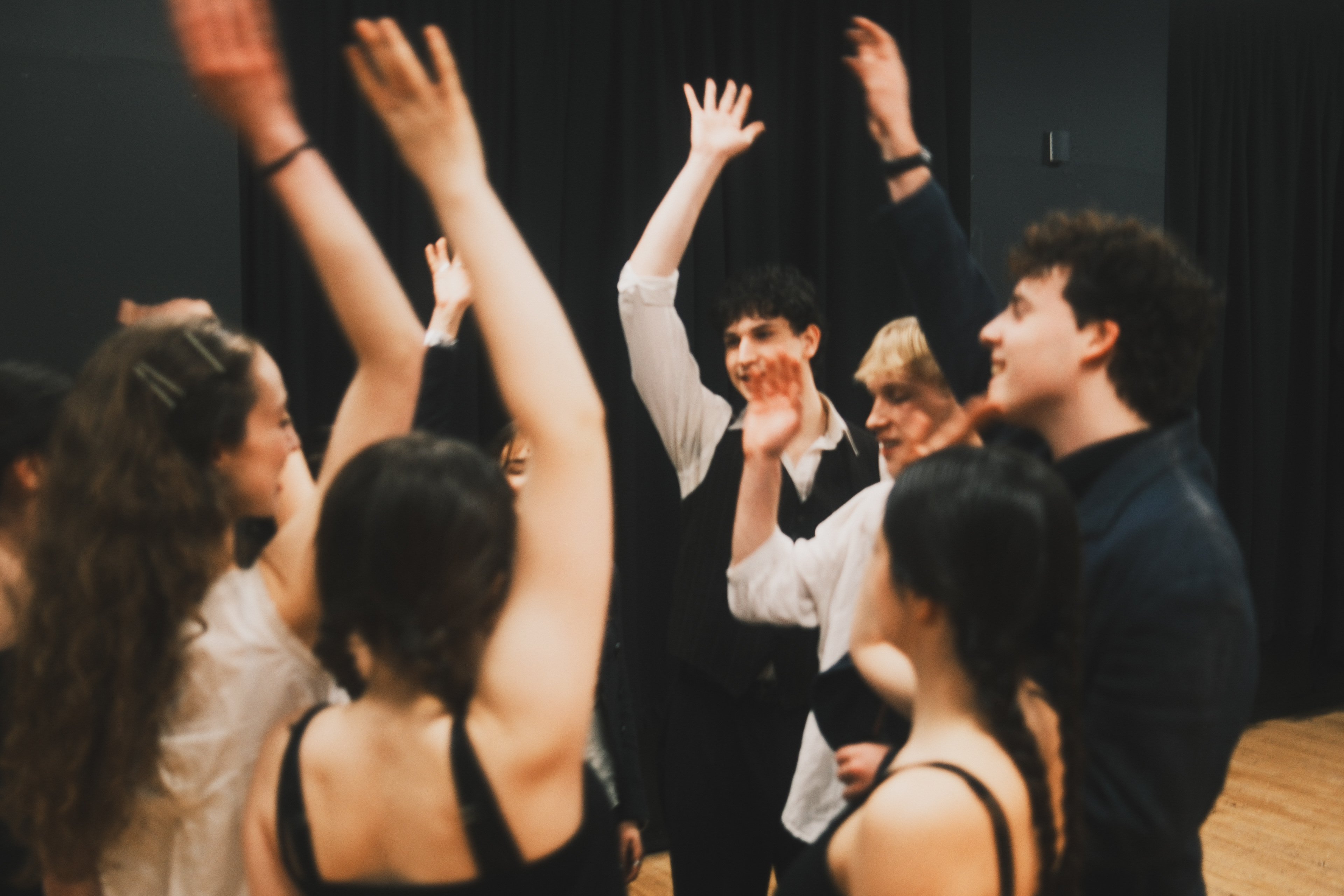 The company in a rehearsal room with hands raised in a circle.