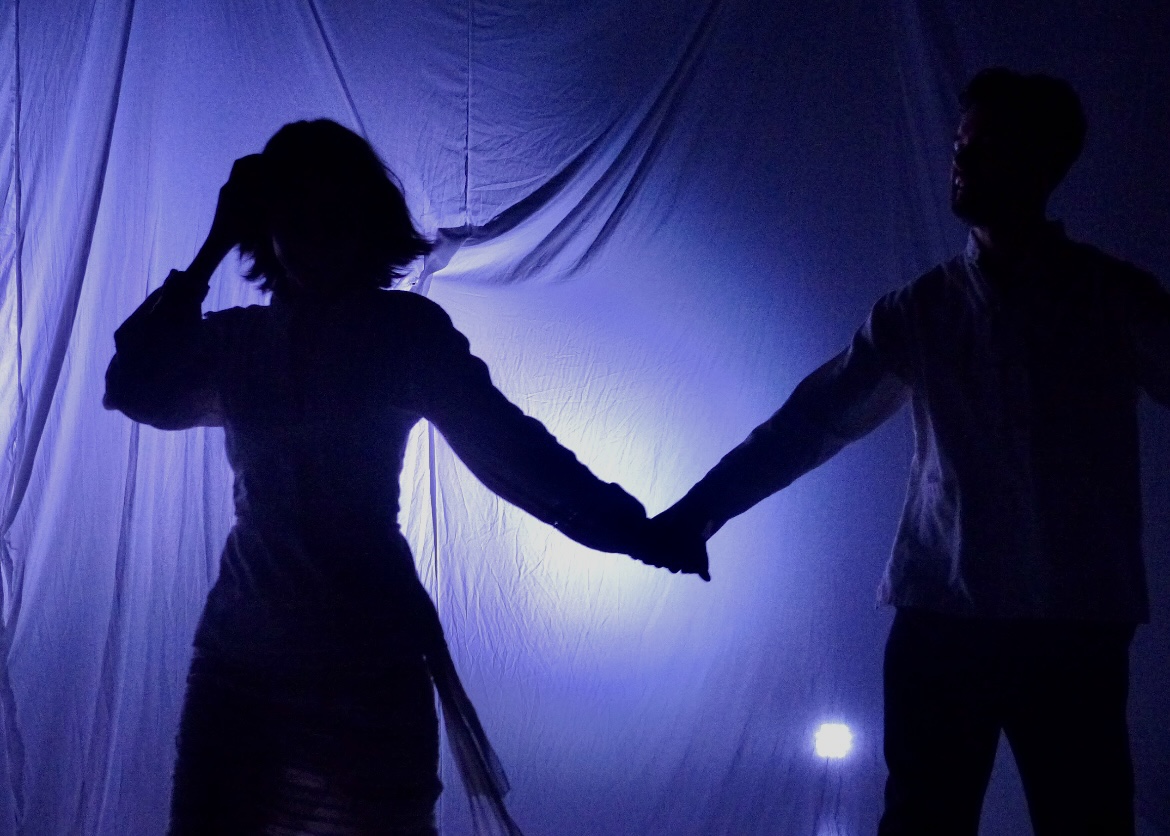 Two silhouetted figures joining hands against a blue-lit sheet.